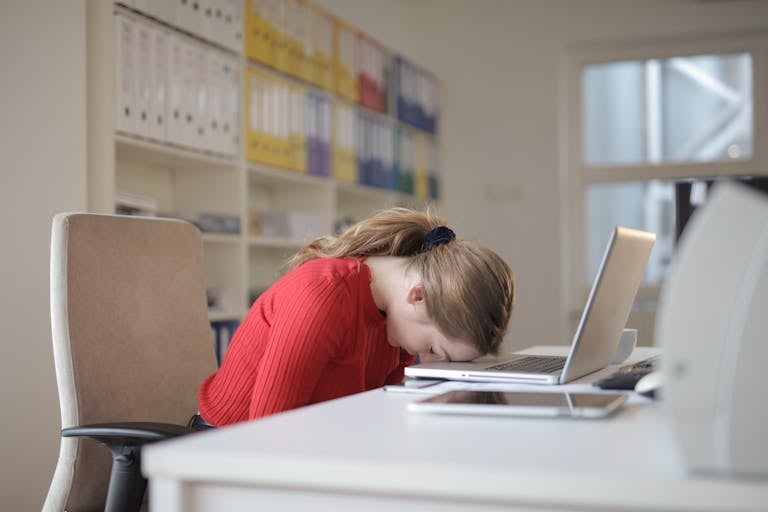A tired adhd woman leans her head on a desk with a laptop, symbolizing workplace fatigue and burnout.