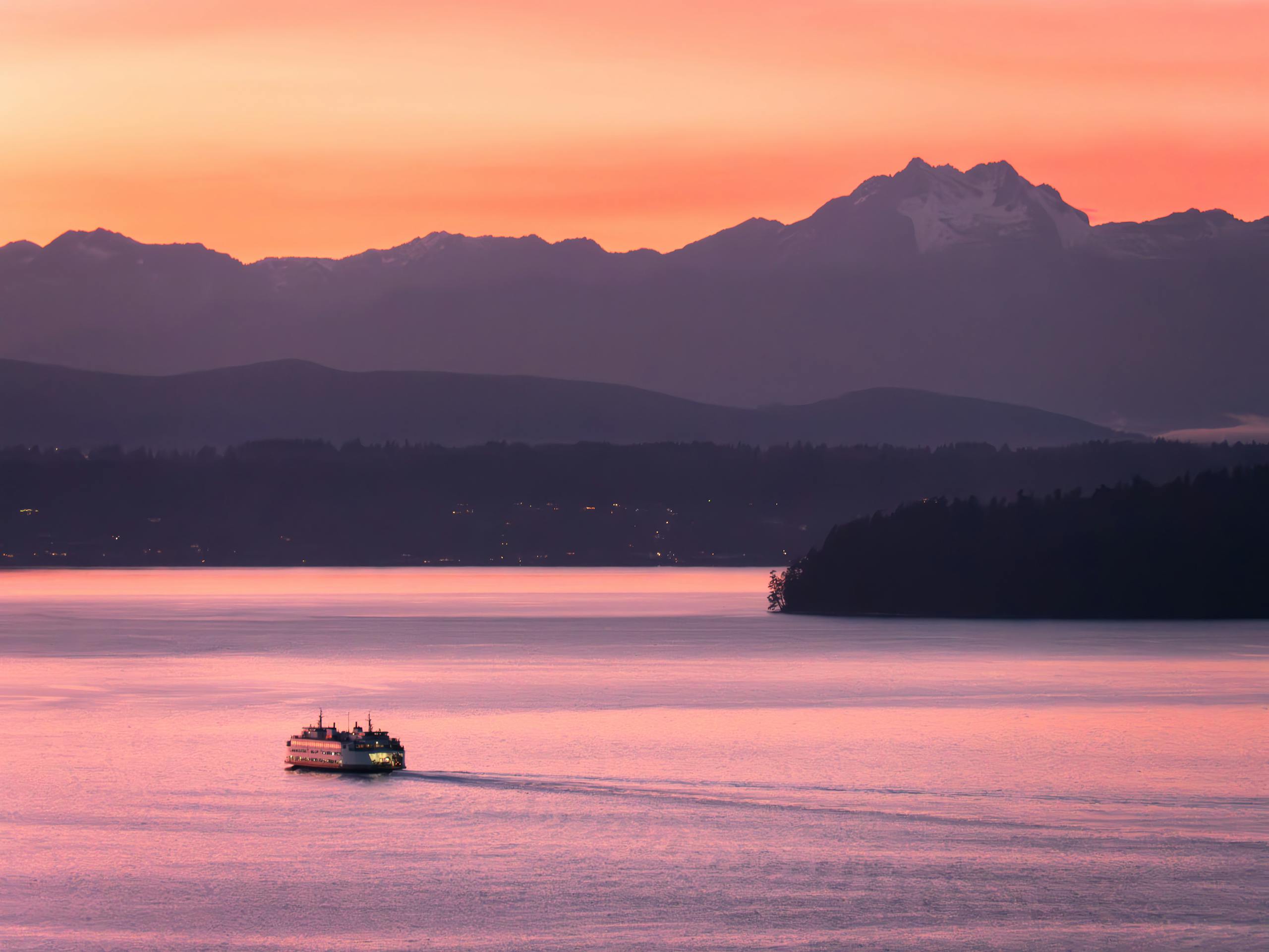 Scenic view of a ferry crossing under a pink sunset sky with mountains in the background in Seattle.